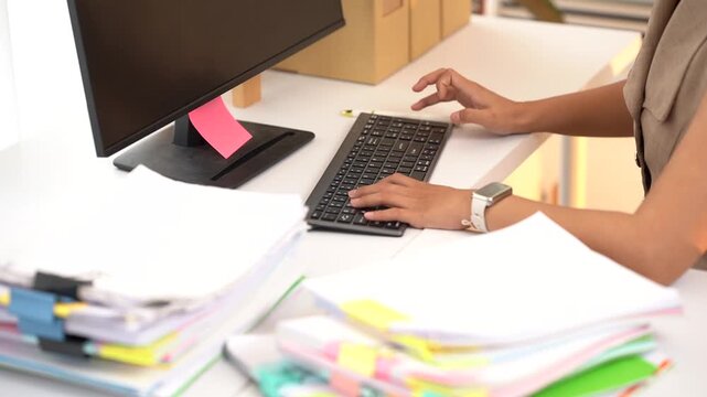 Working diligently at her desk, a professional types on a keyboard, surrounded by stacks of papers and a computer monitor. The scene depicts a focused and productive work environment.