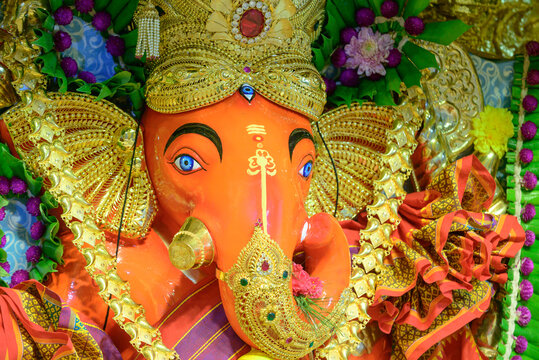 Vibrant orange Ganesha idol adorned with golden ornaments, intricate marigold flowers, and festive purple garlands. Close-up view of traditional Mexican-style religious statue with ornate