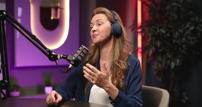 Professional female broadcaster or radio host wearing wireless earphones talking into condenser microphone delivering narrative content during audio podcast session in modern sound recording studio