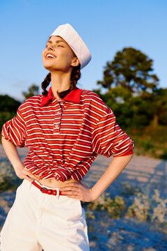 Young woman in striped shirt and hat enjoys sunny outdoor lifestyle during summer vacation. The happy person stands on beach smiling at sun. Concept for travel, leisure, nature, and recreation.