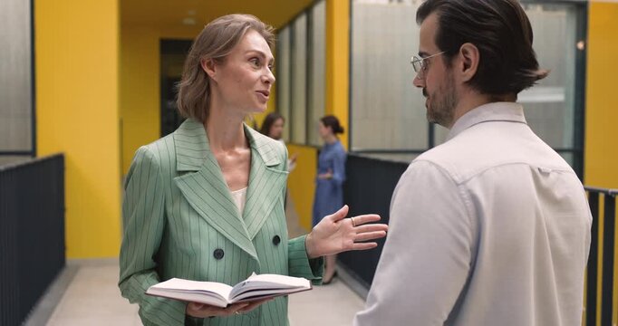 Focused adult businesswoman in formal suit consulting male coworker in atrium office hallway sharing business plan offering solution providing work guidance explaining task referring to paper notebook