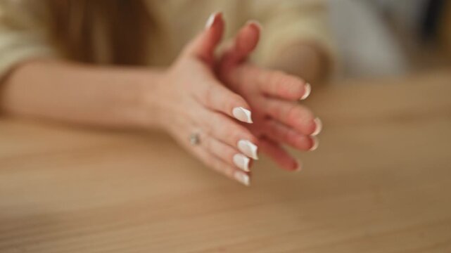 Woman hands clasped on wooden table showing french manicure and ring under soft natural light in studio; thoughtful pause.