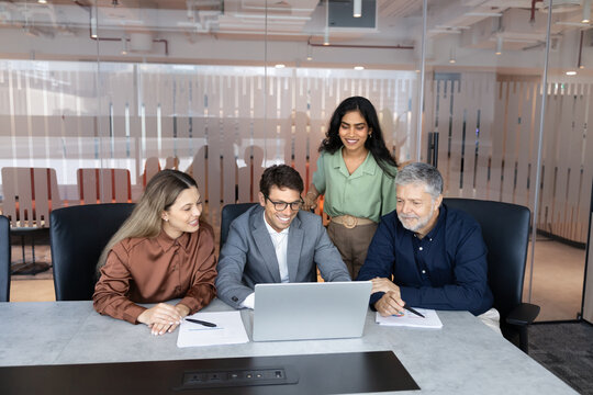 Multiethnic business team gathered around laptop, engaged in discussion during meeting in office conference room. Project management, new corporate platform review, marketing strategy development