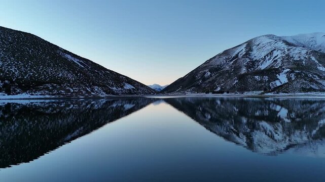 Symmetrical alpine sunrise with mirror reflection on glassy lake between snow-dusted mountains and narrow valley viewpoints from waterline edge, calm blue sky and precise V-shaped composition centered