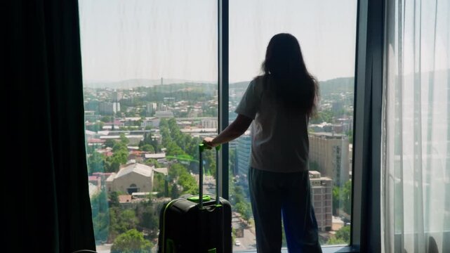 Woman call taxi standing at floor to ceiling window overlooking sprawling city, curtain, rolling luggage nearby, daytime natural light streaming through glass, capturing high rise perspective