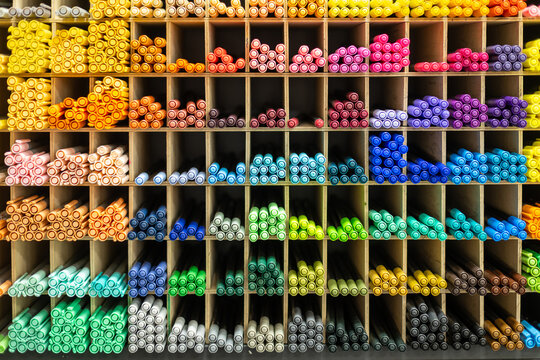 Large assortment of colorful art markers arranged by shades in wooden display shelves at stationery store, vibrant rainbow palette of drawing supplies for artists and designers.