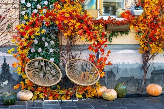 Cozy autumn outdoor seating area with two wicker chairs surrounded by colorful fall foliage, pumpkins and vibrant maple leaves decorating a charming seasonal photo spot.