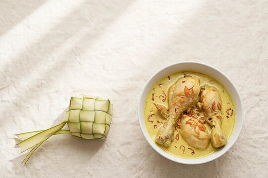 Flat lay of woven ketupat rice cake with bowl of opor ayam coconut chicken curry on cream linen for modern Eid celebration