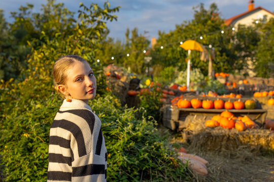 Young blonde girl in a striped sweater looking back at camera in a colorful autumn pumpkin patch, golden sunlight illuminates her face with pumpkins and rustic harvest decorations.