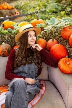 Beautiful teenage girl in a straw hat and burgundy sweater relaxing among colorful pumpkins, thoughtful young brunette enjoying a peaceful autumn day at a rustic harvest festival.