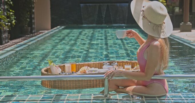 Woman in pink swimsuit and sun hat enjoying floating breakfast in luxury hotel swimming pool. Relaxing morning vacation lifestyle with coffee and tropical food in exotic resort spa.