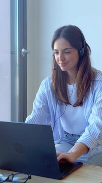 Young smiling woman with a headset working on a laptop computer as a call center agent, providing customer support and assistance while sitting at a desk in her modern home office