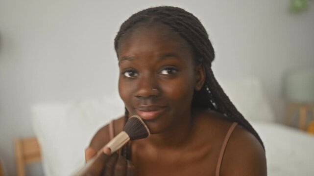 Woman applying makeup with a brush to her face in a studio bedroom setting, holding brush near lips in a closeup beauty routine; confidence.