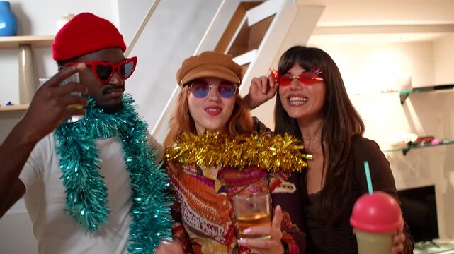 Three diverse and happy friends in funny sunglasses and tinsel boas dancing, drinking, and singing together at a festive house party, celebrating and enjoying a fun filled evening