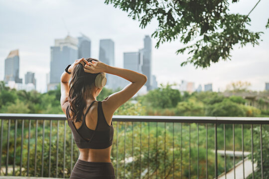 Rear view of young Asian athletic woman tying her hair while wearing wireless headphones and sportswear outdoors, preparing for workout in urban park with green trees