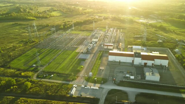 High voltage electrical substation surrounded by green countryside with transformers and pylons distributing energy from the power plant, all illuminated by the warm light of the setting sun