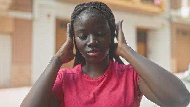 Woman covering ears with hands on a residential street, wearing pink shirt and braided hair; calm reflection.