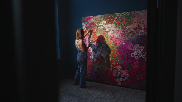 Artist stands before vibrant, oversized floral canvas. Hands brush colors, shaping blossoms in rich hues. Dark room frames the vivid, blooming scene. Creative energy flows through each painted petal