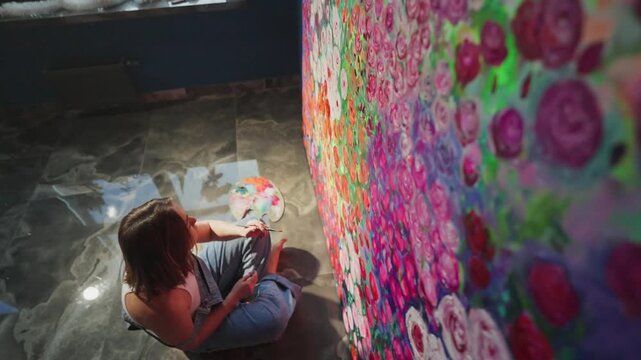 Artist creates vibrant floral mural on large canvas. She sits cross-legged, focused on painting. Soft light glows across the dark tiled floor. Colorful roses bloom in bold, painterly strokes