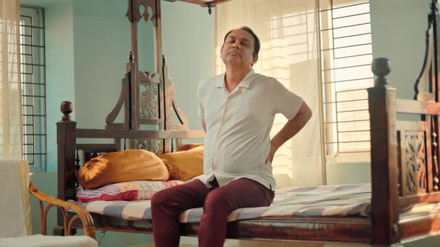 Senior man having lower back pain while sitting on a bed at home, highlighting lumbar pain, muscle strain and spinal discomfort.