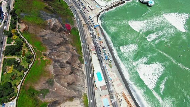 High aerial view of La Rosa Nautica pier in Miraflores, Lima, Peru, with surf breaking on the beach, the Costa Verde highway, cliffs, parks and seaside buildings.