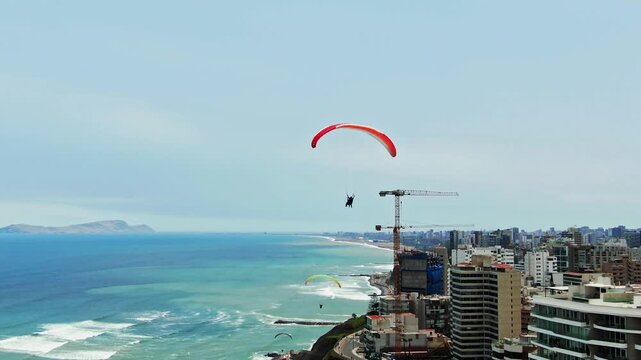 Drone follows a paraglider over the Miraflores coast in Lima, Peru, ending with three paragliders in the sky above the ocean, cliffs, cranes and seaside buildings.