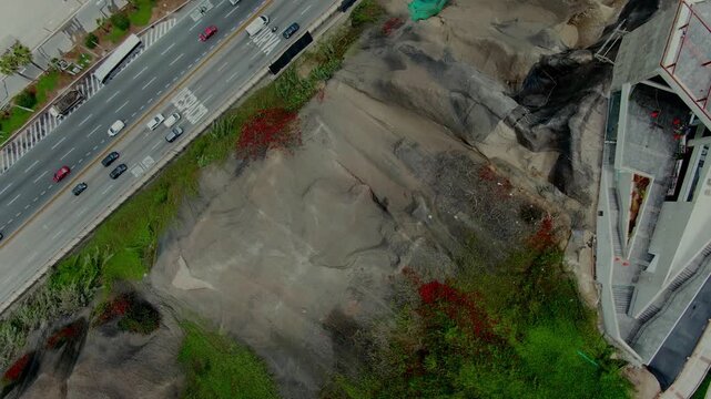 Top-down aerial of the Costa Verde cliff in Miraflores, Lima, Peru, with a burned patch, green erosion mesh, red flowers, the cable car station and highway traffic.