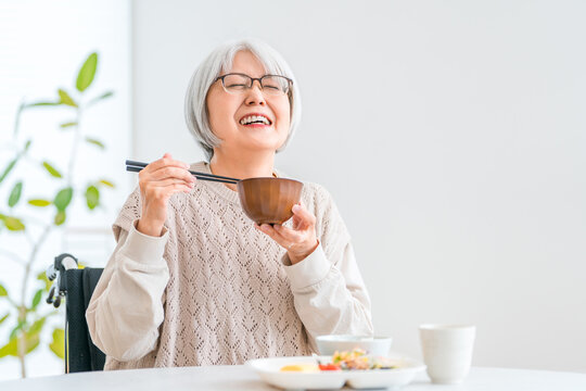 自宅のダイニングで美味しそうにお味噌汁を飲む車椅子ユーザーのシニア女性