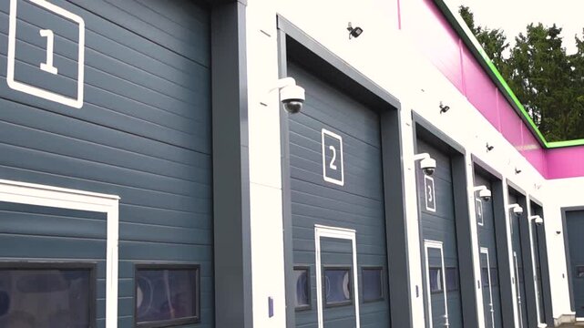 Modern warehouse with numbered gray roll up doors, each equipped with a surveillance camera for enhanced security. The facility features a clean, contemporary design in an urban setting