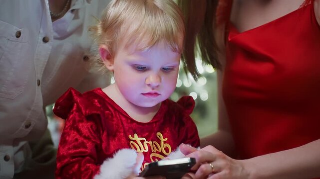 Cute child wears red velvet dress with gold trim. Child holds smartphone with both hands, eyes fixed on screen. Child's face shows focused, slightly curious expression