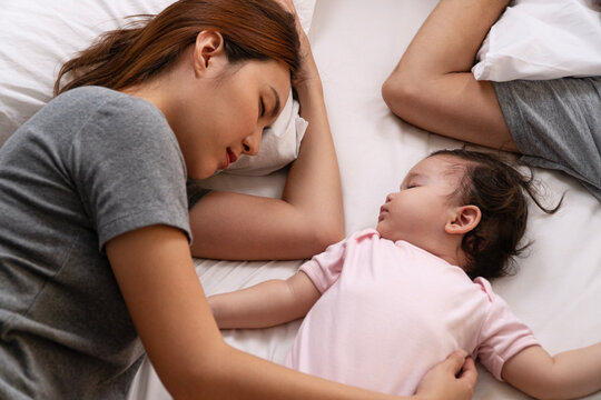 High angle view of a loving young Asian mother and cute baby sleeping peacefully together on a soft white bed at home, representing family bonding, maternity care, and childhood comfort.