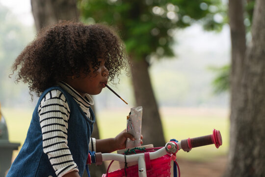 Side profile of a cute little child with curly hair eating a chocolate biscuit stick snack while sitting on a pink bicycle in a green outdoor park, enjoying a happy childhood leisure activity.