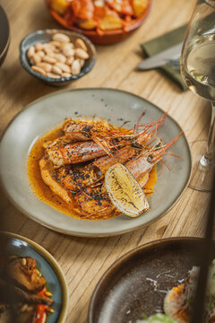 Gourmet grilled prawns served in a rich savory sauce with a grilled lemon wedge on a ceramic plate alongside marcona almonds and a glass of white wine.