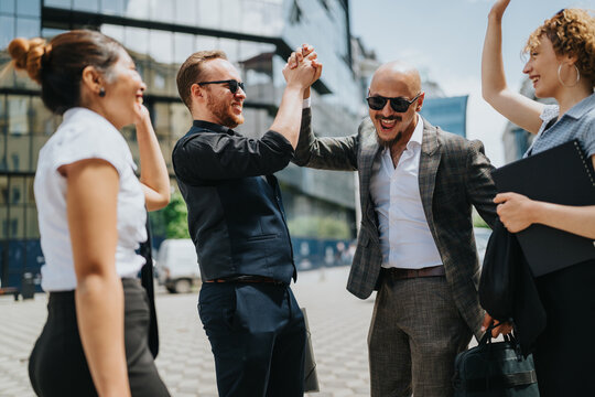 A business team celebrates success outdoors with two men exchanging a high five. Colleagues cheer and laugh together in front of a modern office building during a workday moment.