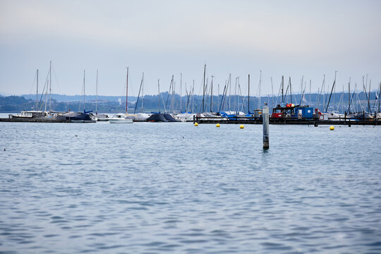 Sailboats moored in a marina, with rows of yachts, dense masts, floating pontoons, and mooring buoys. Recreational boating harbor on a lake or sheltered coastal waterway, with a distant shoreline