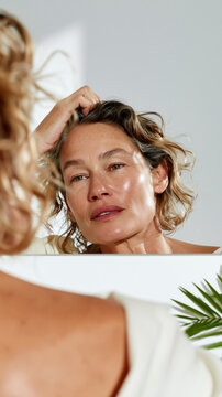 A mature woman gently touches her wavy hair while gazing into a mirror, her natural freckled skin glowing softly during a quiet morning beauty ritual.