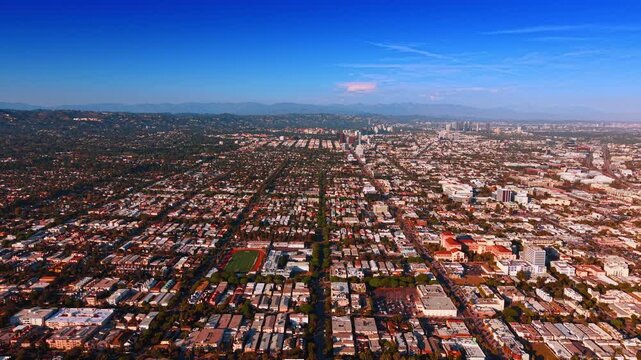 A long avenue divides a city grid beneath distant hills. White roofs flank the central road with trees and a rectangular green field in the foreground.