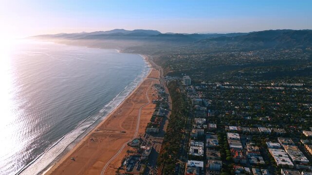 A wide Los Angeles beach follows a bay toward distant mountains. Reflective water fills the left half while paths cross the sand beside tree filled city blocks.
