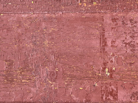 Weathered wooden plank with peeling red paint revealing yellow and brown wood grain and cracked flaking surface texture indicating aged rustic finish and layered brush strokes - close-up view.