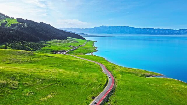 Aerial drone view of a scenic winding highway along the turquoise lake shore and green meadows, Sayram Lake, Xinjiang, China