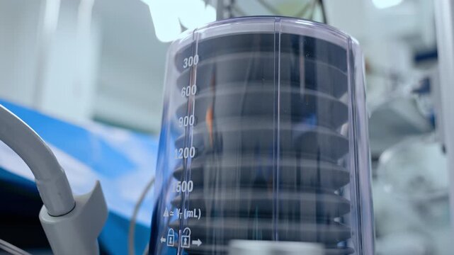 A graduated transparent housing encloses a ribbed medical component. White volume markings run down a cylinder with blue fabric and blurred equipment behind it.
