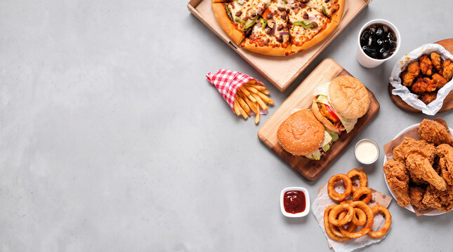 Assortment of fast food with pizza, burgers, fried chicken, and fries with copy space on grey table