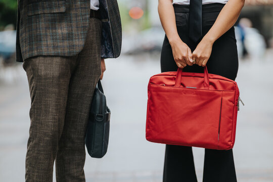 Two business professionals stand side by side holding briefcases at waist level. The scene shows a red laptop bag and a black leather case in an urban commute setting.