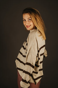 Woman models a striped knit sweater in a studio portrait, smiling over her shoulder against a dark grey background.