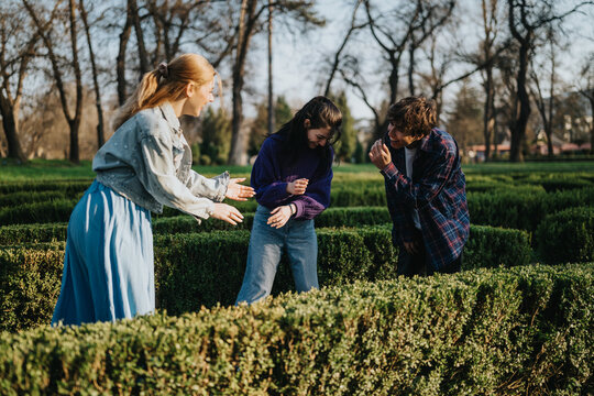 Three friends laugh and interact while standing between trimmed hedges. They enjoy a playful moment together in a park hedge maze on a sunny day.