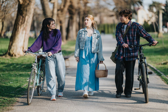 Three friends walk side by side with bicycles and a picnic basket along a tree-lined park path in autumn. They stroll and talk casually, enjoying a relaxed outdoor afternoon together.