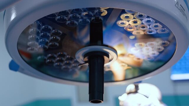 A circular operating lamp fills the foreground above a surgeon. Reflective panels and rows of small lenses occupy the close view against a pale ceiling and blurred cap.
