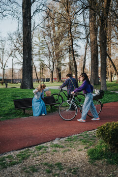 Three friends with bicycles spend time together in a city park near a bench. Two women and a man walk bikes past a seated woman holding a basket among tall trees.