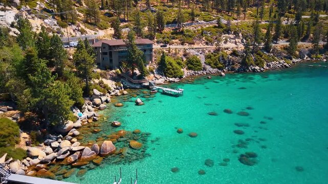 A lakeside house overlooks boulders and turquoise shallows. Boats rest near the pine covered shore while submerged stones form dark patches beneath the water.