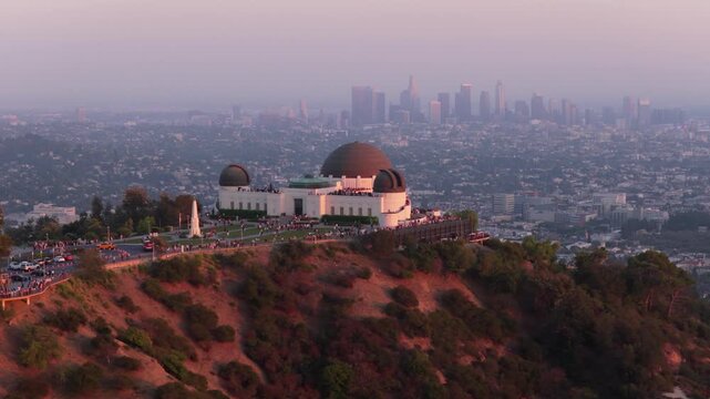 Griffith Observatory with copper domes crowds terraces on a hilltop over Los Angeles, California. Aerial view of downtown skyline in hazy sunset light, cars on road, dry brush slope and green trees.